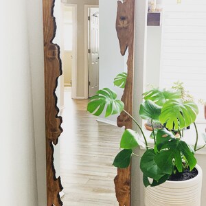 May include: A large, rectangular mirror with a unique, natural wood frame. The frame has a dark brown stain and a rough, textured edge. The mirror is reflecting a room with hardwood floors and a plant in a white pot.