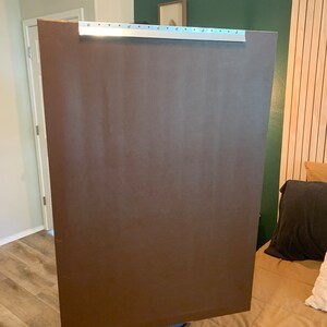 May include: A person holding a large, brown, rectangular piece of cardboard with a silver metal strip along the top edge.