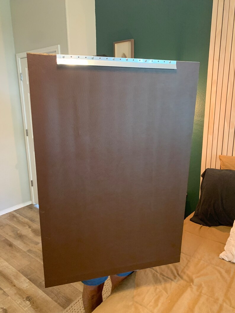 May include: A person holding a large, brown, rectangular piece of cardboard with a silver metal strip along the top edge.