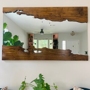 May include: A large, rectangular mirror with a unique, natural wood frame. The frame has a live edge design with a dark brown stain. The mirror reflects a room with a ceiling fan and a door.