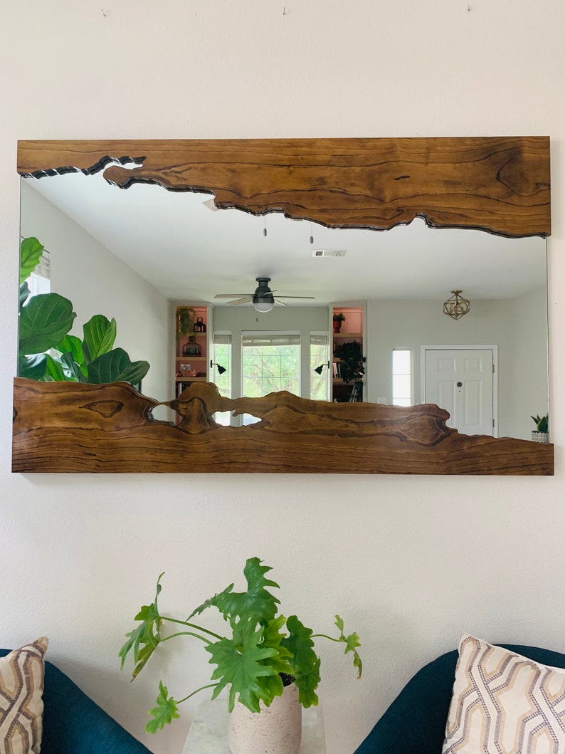 May include: A large, rectangular mirror with a unique, natural wood frame. The frame has a live edge design with a dark brown stain. The mirror reflects a room with a ceiling fan and a door.