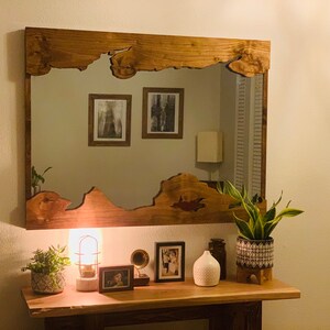 May include: A wooden mirror with a unique, irregular shape frame. The mirror is hanging on a wall above a wooden console table with a lamp, plants, and framed photos.