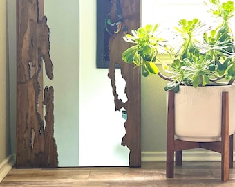 May include: A large, rectangular mirror with a wooden frame. The frame has a natural, organic edge, with a dark brown color. The mirror's surface is divided into sections, with a clear reflective surface. The mirror is leaning against a wall.