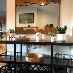 May include: A wooden framed mirror with a unique, natural edge design. The mirror is hanging above a black metal bar cart with two shelves. The bar cart has a wooden top and a wine rack. The mirror is 38 inches by 50 inches.
