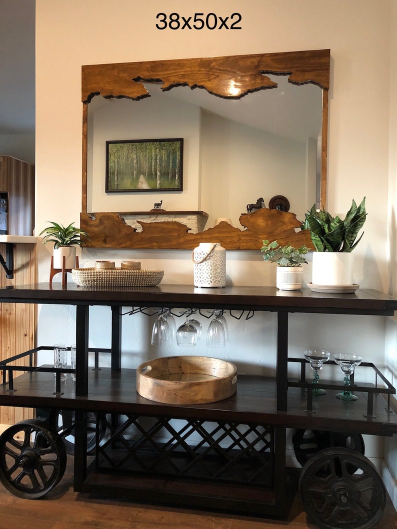May include: A wooden framed mirror with a unique, natural edge design. The mirror is hanging above a black metal bar cart with two shelves. The bar cart has a wooden top and a wine rack. The mirror is 38 inches by 50 inches.