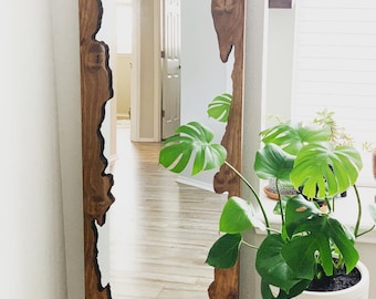 May include: A full-length mirror with a unique, natural wood frame. The frame has a live edge design with a dark brown stain. The mirror is reflecting a room with hardwood floors and a plant in a white pot.