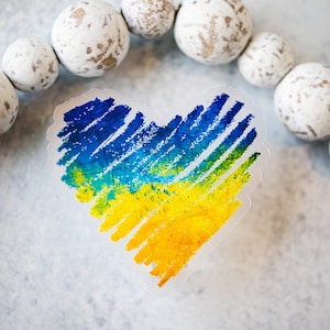 May include: A heart-shaped sticker with a blue and yellow design, reminiscent of the Ukrainian flag. The sticker is placed on a white surface with a string of white beads around it.
