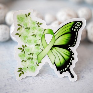 K&ouml;nnte beinhalten: Ein schmetterlingsf&ouml;rmiger Aufkleber mit einem gr&uuml;nen Band und floralem Design. Die linke Seite zeigt hellgr&uuml;ne Rosen und Bl&auml;tter, w&auml;hrend die rechte Seite einen gr&uuml;nen und schwarzen Schmetterlingsfl&uuml;gel zeigt. Der Aufkleber befindet sich auf einer hellblauen Oberfl&auml;che.