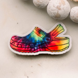 Puede incluir: Una pegatina de zapato Crocs con estampado tie-dye. La pegatina es blanca con un diseño tie-dye colorido en el zapato.
