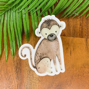 May include: A brown and white monkey sticker with a curled tail. The monkey is sitting with its legs crossed and has a cute, cartoon-like expression.