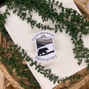 Puede incluir: Una pegatina blanca con una silueta de oso negro caminando sobre una cordillera con una salida del sol en el fondo. El texto "The Great Smoky Mountains National Park" est&aacute; impreso en la pegatina.