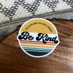May include: A round sticker with a retro design featuring a yellow, orange, and green striped background. The sticker says "In a world where you can be anything, Be Kind" in black text.