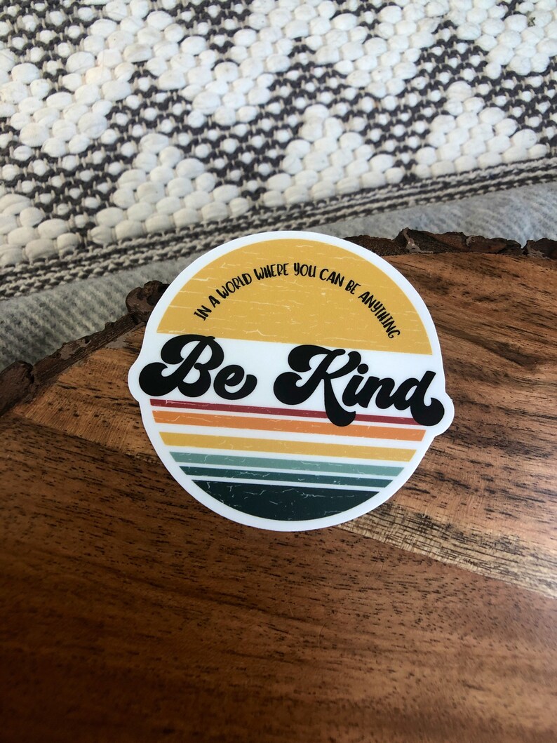 May include: A round sticker with a retro design featuring a yellow, orange, and green striped background. The sticker says "In a world where you can be anything, Be Kind" in black text.