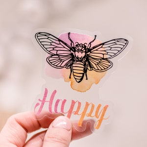 May include: A clear sticker featuring a black line drawing of an insect with detailed wings, set against a watercolor wash of pink, orange, and yellow. The word "Happy" is written in pink script below the insect.