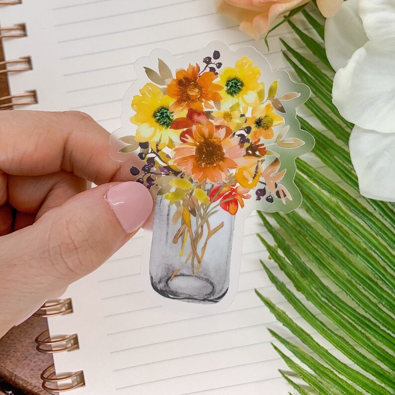 Puede incluir: Un jarr&oacute;n de vidrio transparente lleno de un ramo de girasoles, margaritas y otras flores de oto&ntilde;o en acuarela en tonos amarillos, naranjas y rojos.