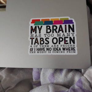 May include: A white sticker with a colorful row of tabs at the top. The sticker says "MY BRAIN HAS TOO MANY TABS OPEN 4 OF THEM ARE FROZEN & I HAVE NO IDEA WHERE THE MUSIC IS COMING FROM".