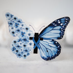 May include: A blue butterfly with a cross and ribbon on its body. The butterfly is made of clear plastic and has a floral design on one wing.