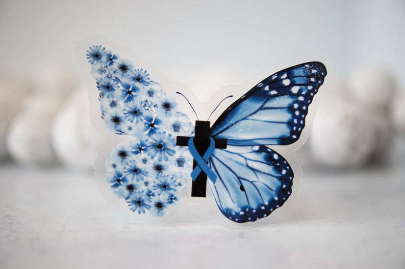 May include: A blue butterfly with a cross and ribbon on its body. The butterfly is made of clear plastic and has a floral design on one wing.