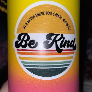 May include: A yellow and pink water bottle with a white sticker that says "In a world where you can be anything, Be Kind". The sticker has a retro design with a sunburst and stripes.