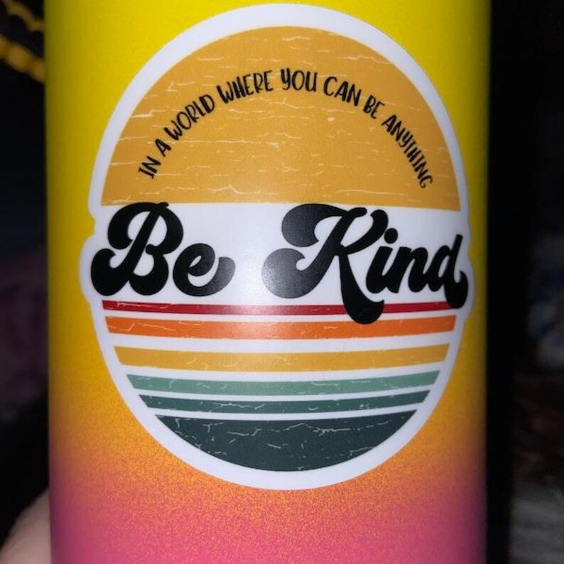 May include: A yellow and pink water bottle with a white sticker that says "In a world where you can be anything, Be Kind". The sticker has a retro design with a sunburst and stripes.
