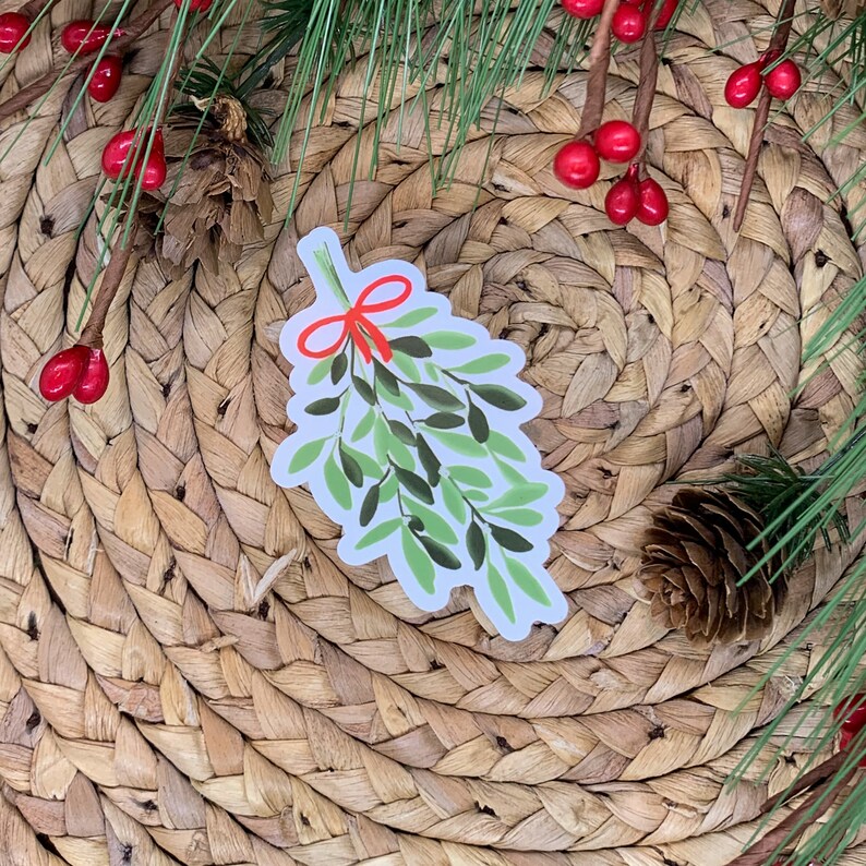 May include: A white sticker with a green and red illustration of a sprig of mistletoe with a red bow. The sticker is on a woven brown background.