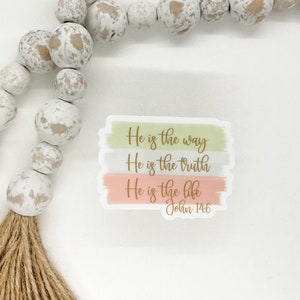 Pode incluir: Um adesivo com um fundo de aquarela em tons de verde, cinza e rosa. O adesivo apresenta o texto "Ele &eacute; o caminho, Ele &eacute; a verdade, Ele &eacute; a vida" em letras douradas. O texto "Jo&atilde;o 14:6" tamb&eacute;m est&aacute; inclu&iacute;do em letras douradas.