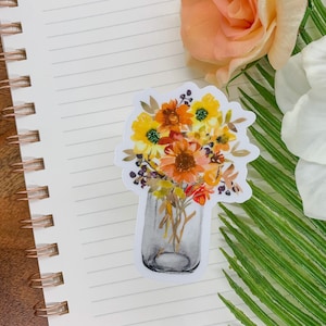 Puede incluir: Una pegatina con una ilustraci&oacute;n de acuarela de un tarro de cristal lleno de un ramo de flores amarillas, naranjas y rojas. Las flores est&aacute;n dispuestas en un estilo r&uacute;stico, con algunos tallos que sobresalen del tarro.
