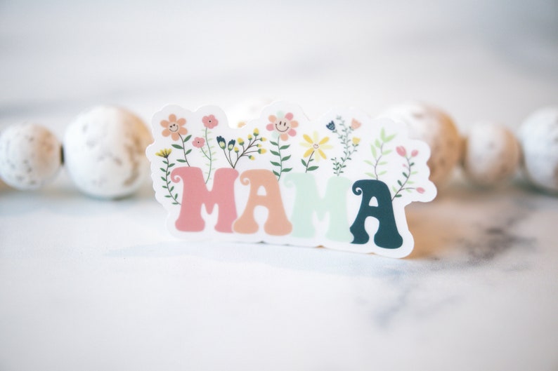 Pode incluir: Um adesivo colorido com a palavra "MAMA" em uma fonte caprichosa, cercada por flores e folhas.