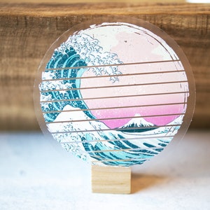 May include: A round, clear acrylic coaster with a printed image of a blue and white wave crashing over a pink and white mountain. The coaster is on a wooden stand.