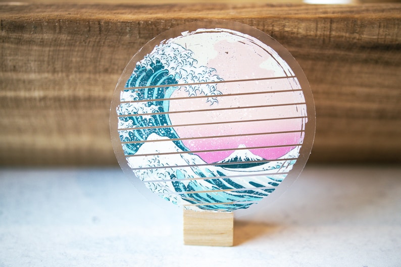 May include: A round, clear acrylic coaster with a printed image of a blue and white wave crashing over a pink and white mountain. The coaster is on a wooden stand.