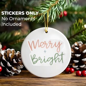 K&ouml;nnte beinhalten: Wei&szlig;er Keramik-Anh&auml;nger mit dem Text "Merry + Bright" in rosa und gr&uuml;ner Schrift. Der Anh&auml;nger h&auml;ngt an einem goldenen Band und ist von Kiefern&auml;sten und roten Beeren umgeben.