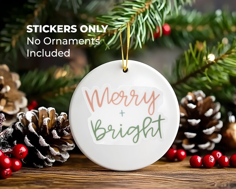 K&ouml;nnte beinhalten: Wei&szlig;er Keramik-Anh&auml;nger mit dem Text "Merry + Bright" in rosa und gr&uuml;ner Schrift. Der Anh&auml;nger h&auml;ngt an einem goldenen Band und ist von Kiefern&auml;sten und roten Beeren umgeben.