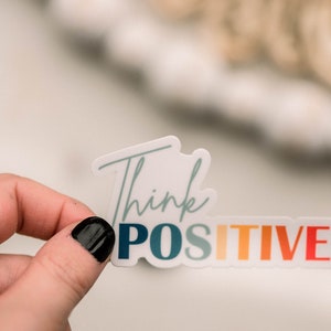 May include: A white sticker with the text "Think Positive" in a rainbow gradient. The sticker is being held by a hand with black nail polish.