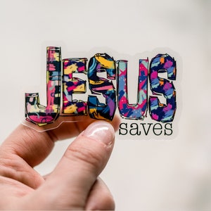 May include: A clear sticker with the word "JESUS" in colorful, abstract lettering and the word "saves" in black lettering below.