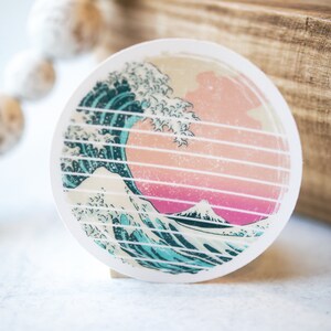 May include: A round sticker with a graphic of a blue and white wave crashing over a pink and white mountain. The wave is stylized to look like a traditional Japanese woodblock print.