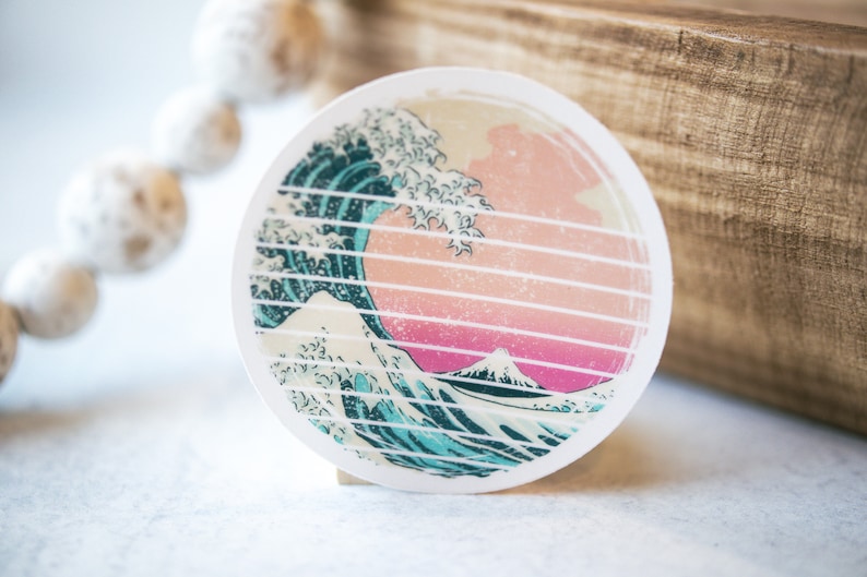 May include: A round sticker with a graphic of a blue and white wave crashing over a pink and white mountain. The wave is stylized to look like a traditional Japanese woodblock print.