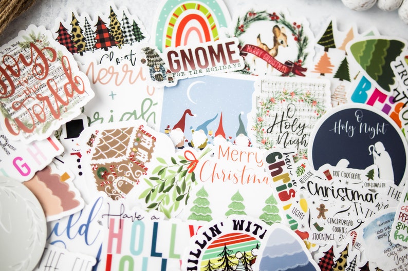 K&ouml;nnte beinhalten: Eine Auswahl an weihnachtlichen Aufklebern mit verschiedenen Designs, darunter ein Lebkuchenhaus, ein Gnom, ein Rentier und ein Weihnachtsbaum. Die Aufkleber sind auf wei&szlig;em Papier mit bunten Designs und Text gedruckt.