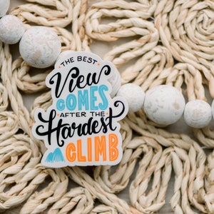 May include: White sticker with blue and orange text that reads "The best view comes after the hardest climb". The sticker is on a woven straw surface with white beads.