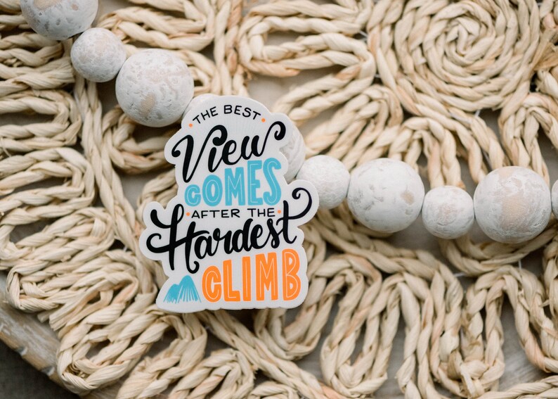 May include: White sticker with blue and orange text that reads "The best view comes after the hardest climb". The sticker is on a woven straw surface with white beads.