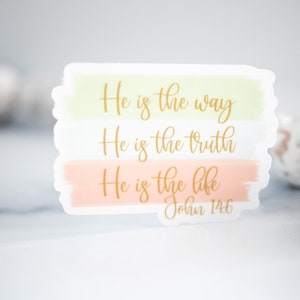 Pode incluir: Um adesivo branco com letras douradas que diz "Ele &eacute; o caminho, Ele &eacute; a verdade, Ele &eacute; a vida, Jo&atilde;o 14:6". O adesivo tem um fundo de aquarela com cores verde claro, branco e p&ecirc;ssego.