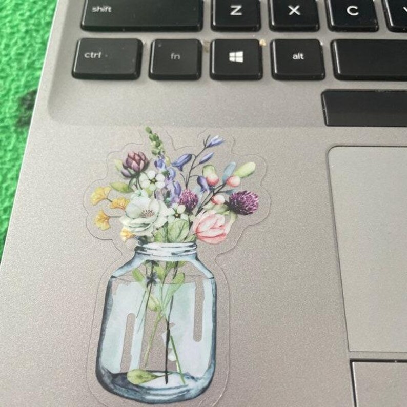 May include: A clear glass jar with a bouquet of colorful wildflowers, including purple, pink, white, and yellow flowers. The jar is sitting on a silver surface.