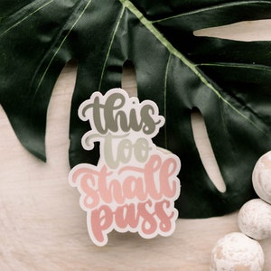 May include: A white sticker with the text "This too shall pass" in pink, green, and gray lettering. The sticker is on a light wood surface with a green leaf.