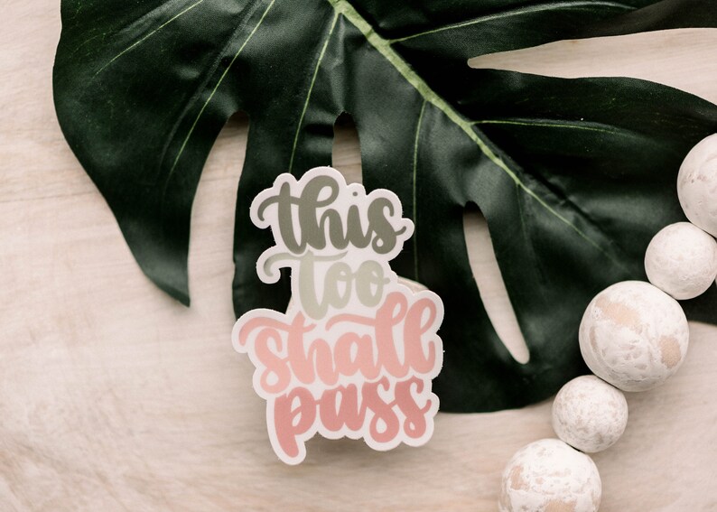 May include: A white sticker with the text "This too shall pass" in pink, green, and gray lettering. The sticker is on a light wood surface with a green leaf.