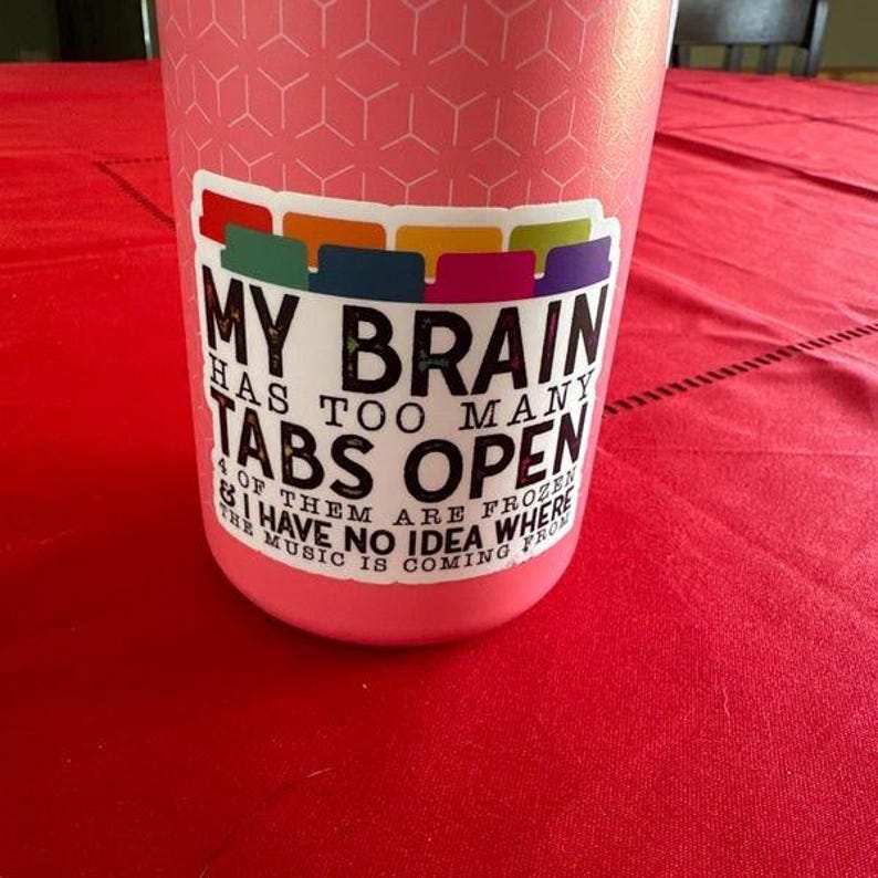 May include: A pink insulated tumbler with a white sticker that says "My brain has too many tabs open & of them are frozen & I have no idea where the music is coming from". The sticker has a row of colorful tabs at the top.