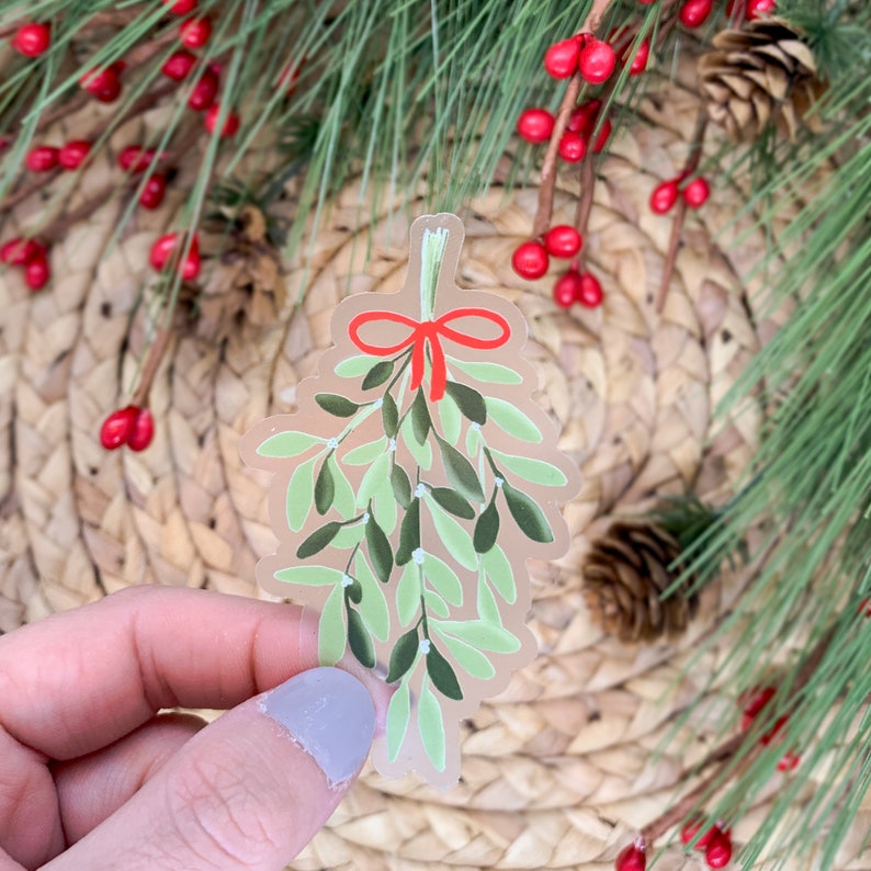 May include: A clear sticker of a green mistletoe sprig with a red bow. The mistletoe is tied with a red ribbon and has white berries.