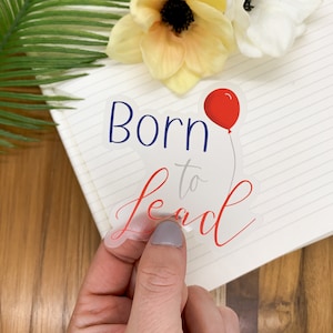 Op de afbeelding: Een transparante sticker met de tekst "Born to Teach" in blauwe en rode letters. Een rode ballon bevindt zich boven de tekst.