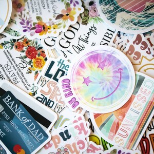 May include: A collection of colorful stickers with various designs and text. Some stickers feature floral arrangements, inspirational quotes, and a tie-dye smiley face. One sticker reads "BANK of DAD" with a credit card design.