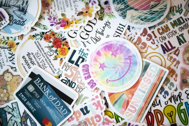 May include: A collection of colorful stickers with various designs and text. Some stickers feature floral arrangements, inspirational quotes, and a tie-dye smiley face. One sticker reads "BANK of DAD" with a credit card design.