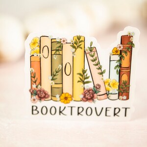 Pu&ograve; includere: Un adesivo con una pila di libri con accenti floreali e il testo "BOOKTROVERT".