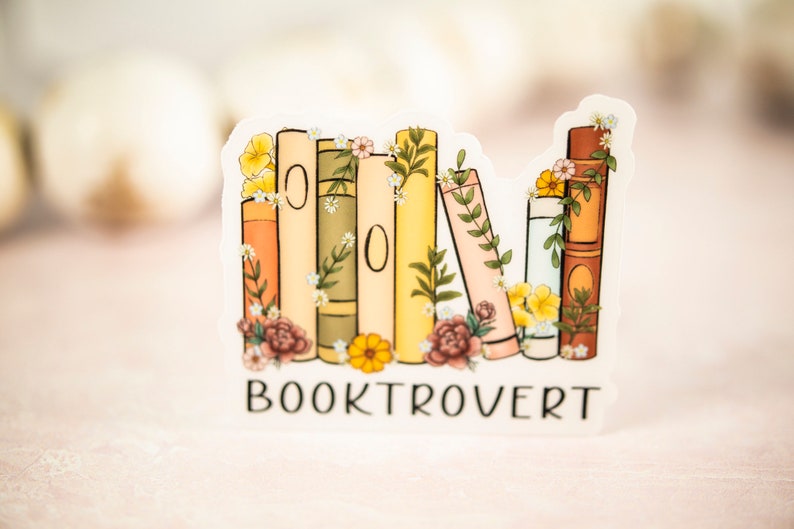 Pu&ograve; includere: Un adesivo con una pila di libri con accenti floreali e il testo "BOOKTROVERT".